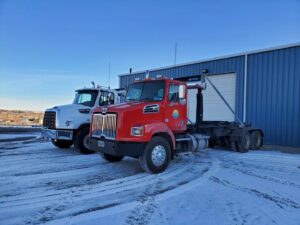 Two roll-off trucks, one white and one red, parked in a snowy lot for First Light Services Llc in Casper, WY.