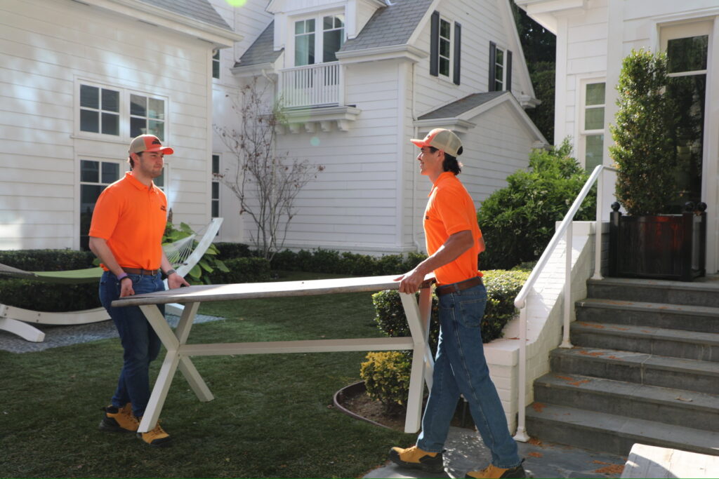 Two The Junkiez Junk Removal workers carrying a long table across a residential lawn in Los Angeles, CA.
