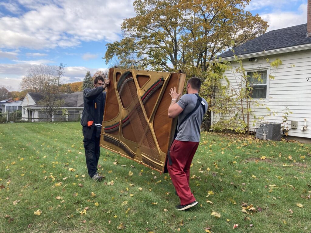 Two men carrying a heavy piano soundboard across a yard, a service provided by Hometown Hauling 802 in Milton, VT.