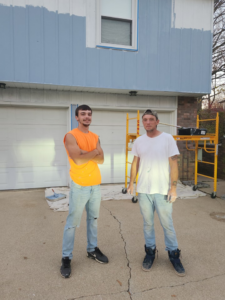 Two men standing in front of a house with partially painted blue and white siding, showing exterior painting work by Drew's Handy Construction in Charleston, IL.