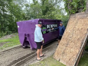 Two men standing next to a DumpDog purple dumpster at a cleanup site, ready for junk removal in Flint, MI.
