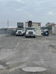 Two roll-off trucks, one with a container and another with its lifting arm raised, near a dumpster at A-1 Roll-Off Rentals, LLC in Baton Rouge, LA.