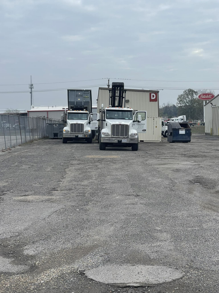 Two roll-off trucks, one with a container and another with its lifting arm raised, near a dumpster at A-1 Roll-Off Rentals, LLC in Baton Rouge, LA.