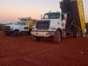 Two roll-off trucks, one with a yellow dumpster, parked on a dirt job site for Tecumseh Rolloff Services in Moore, OK.