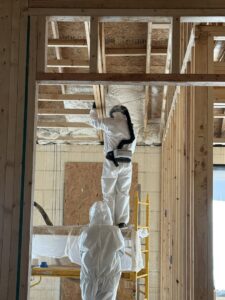 Two workers in protective suits applying spray foam insulation to walls and ceiling for Off-Axis Spray Foam and Radon Services in Clarksville, TN.
