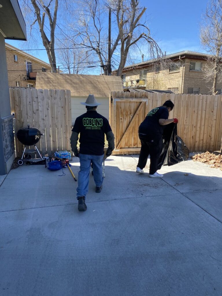Two Goblins Junk Removal workers, one carrying a trash bag, actively cleaning up a backyard patio area in Wheat Ridge, CO.