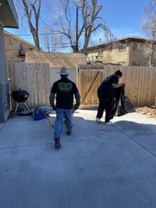 Two Goblins Junk Removal workers cleaning up a backyard patio, with one carrying a large trash bag, in Wheat Ridge, CO.