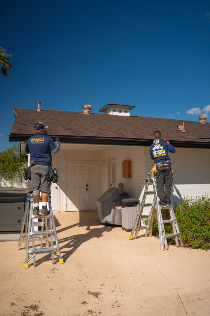 Two workers from Phoenix Rain Gutters and Sun Protection on ladders installing new rain gutters on a house in Glendale, AZ.