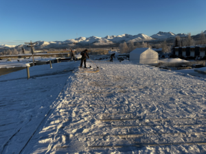 Two workers from AK Property Maintenance, Inc. are shoveling snow off a large flat roof in Anchorage, AK.