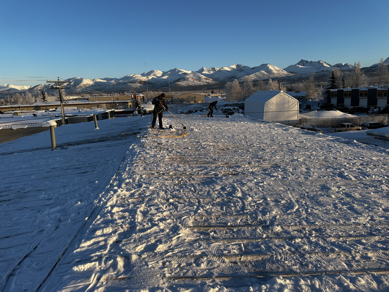 Two workers from AK Property Maintenance, Inc. are shoveling snow off a large flat roof in Anchorage, AK.