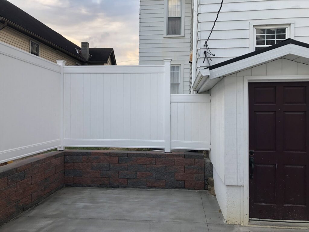 A newly installed white vinyl fence, paver retaining wall, and concrete patio by Gold-Mine Construction, LLC in Pittsburgh, PA.