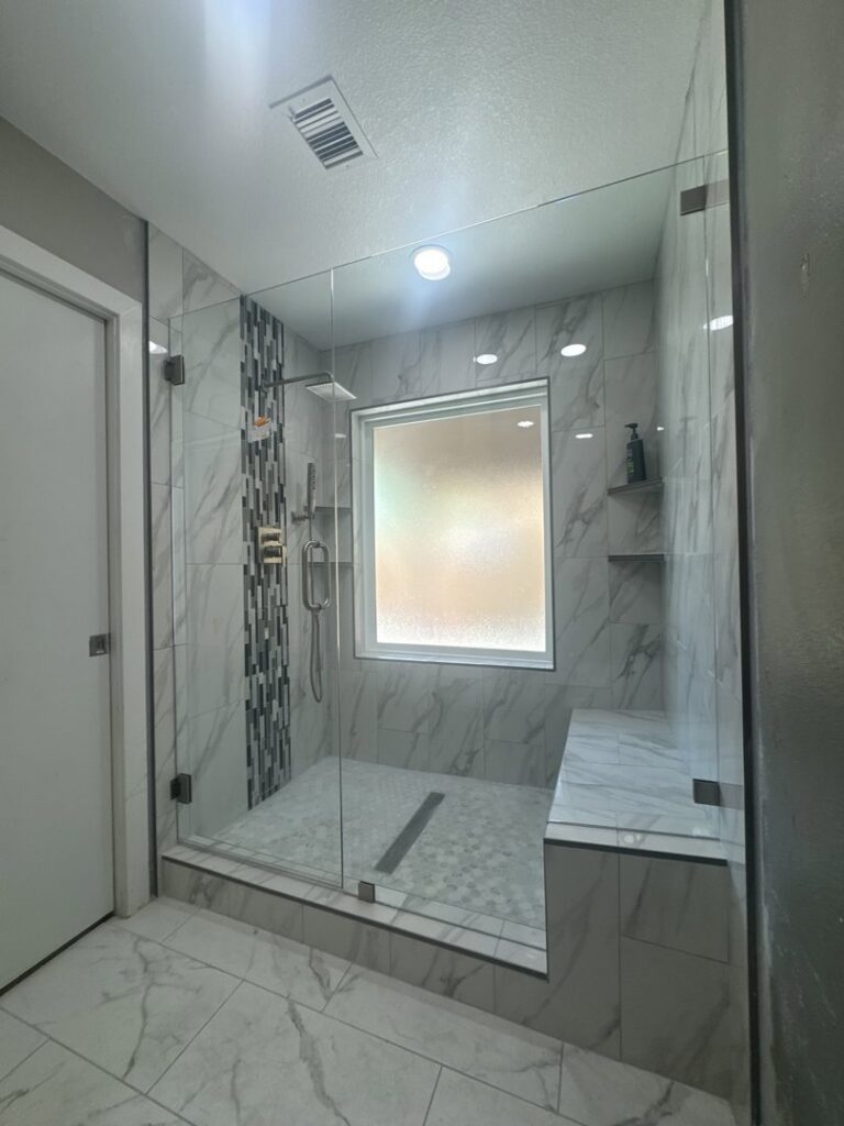 A newly installed walk-in shower with glass doors, marble-look tiles, and a built-in bench by American Wolf Construction in Mesa, AZ