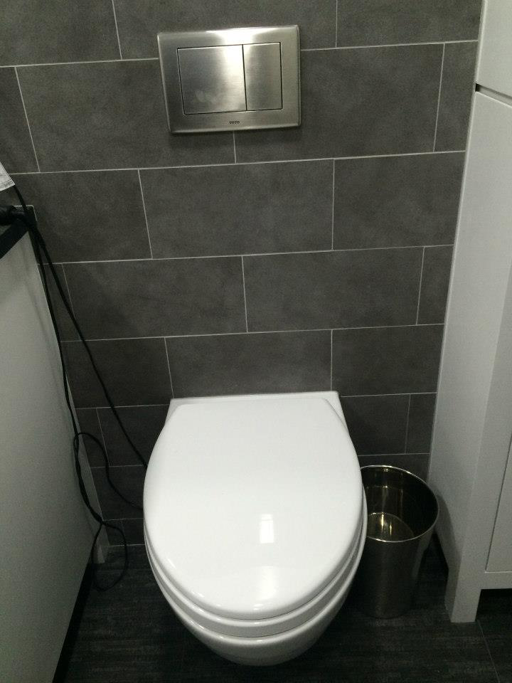 A modern wall-mounted toilet with a flush plate and dark tiled walls in a bathroom by Executech Interiors in Yonkers, NY