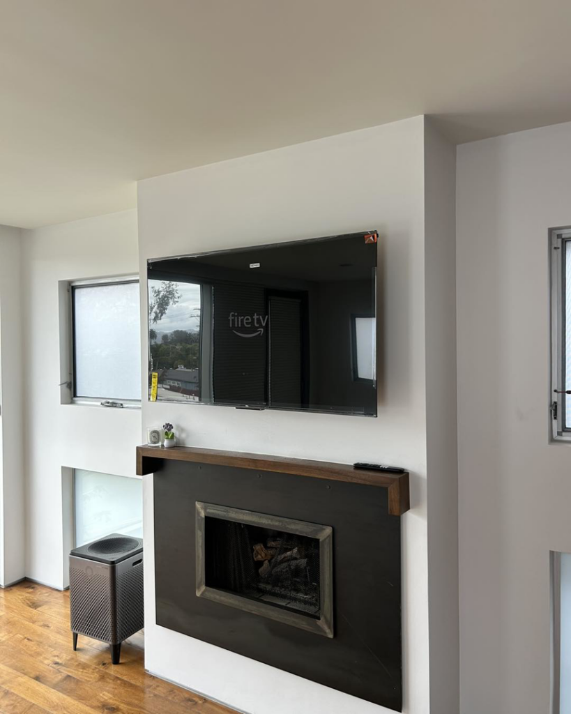 A professionally installed wall-mounted TV above a modern fireplace with a wooden mantel by In&Out Handyman in West Hollywood, CA.