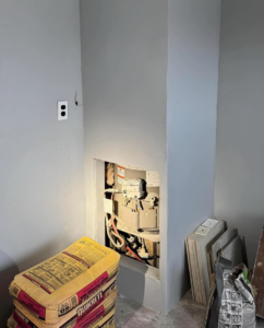 A wall opening revealing plumbing or a meter, with bags of Quikrete cement mix, indicating a renovation project by The ABQ Handyman in Albuquerque, NM.