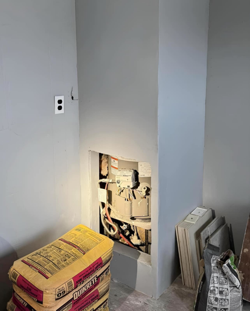 A wall opening revealing plumbing or a meter, with bags of Quikrete cement mix, indicating a renovation project by The ABQ Handyman in Albuquerque, NM.
