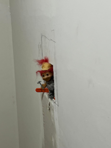 Troll doll with hard hat and hammer peeking from a wall hole, showing wall repair by Dove Construction and Remodeling in Springfield, MO.