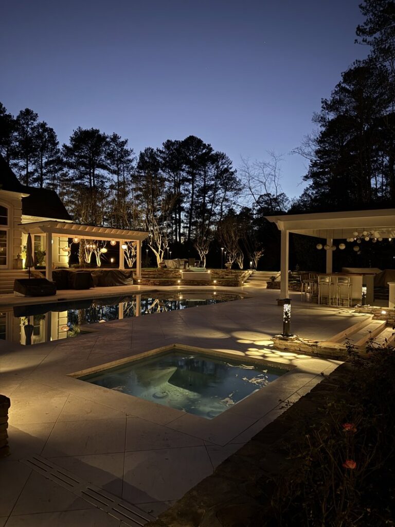 A backyard pool and spa area beautifully illuminated with warm ambient lighting at night by Picasso Poolscapes in Johns Creek, GA.