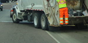 A waste collection worker in safety gear on the back of a garbage truck for Patriot Sanitation Management in Raleigh, NC
