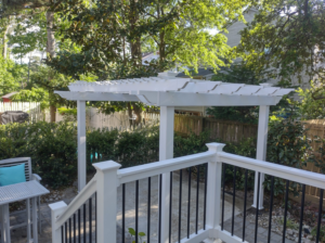 A white pergola in a lush backyard, viewed from a deck, by Signature Deck & Pergola in Virginia Beach, VA