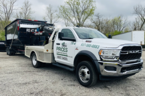 A white RAM truck from Prices Dumpster Rental towing a roll-off dumpster filled with junk for general removal in Dayton, OH.