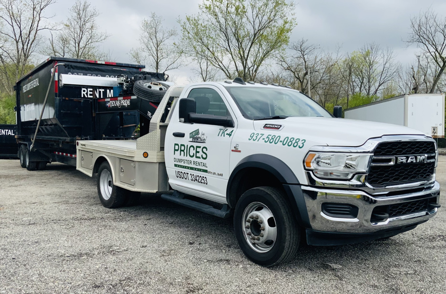 A white RAM truck from Prices Dumpster Rental towing a roll-off dumpster filled with junk for general removal in Dayton, OH.
