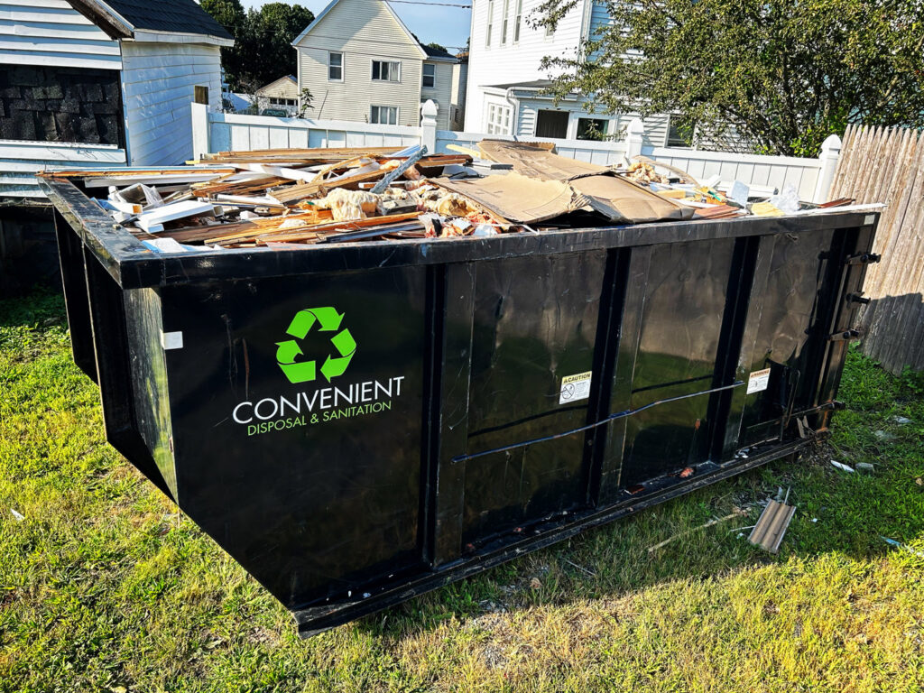 A white roll-off truck delivering a black dumpster to a residential driveway for junk removal by Convenient Disposal & Sanitation in Fall River, MA.