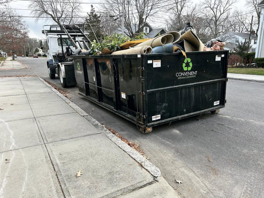 A white roll-off truck picking up a black dumpster from a residential driveway after a junk removal job by Convenient Disposal & Sanitation in Fall River, MA.