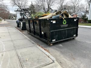 A white roll-off truck picking up a black dumpster from a residential driveway after a junk removal job by Convenient Disposal & Sanitation in Fall River, MA.