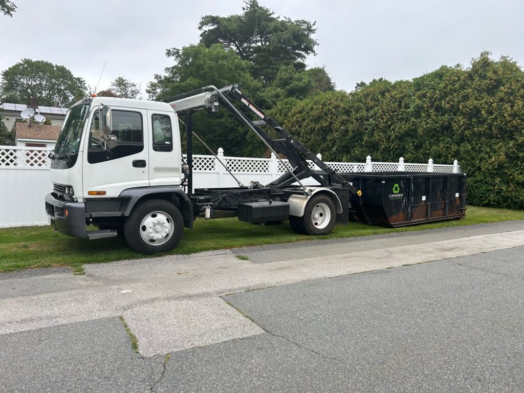A white roll-off truck delivering a black dumpster to a residential street for junk removal by Convenient Disposal & Sanitation in Fall River, MA.