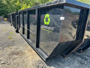 A white roll-off truck hauling a full black dumpster with junk and debris on a residential street in Fall River, MA.