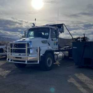 A white roll-off truck parked next to a large dumpster, showcasing junk removal services by First Light Services Llc in Casper, WY.