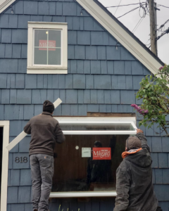 Two workers installing a new window on the exterior of a blue shingle house by RG Construction WA, LLC in Portland, OR.