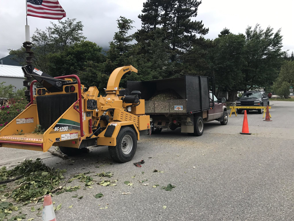 A yellow Vermeer wood chipper and dump truck on a job site, with tree branches on the ground, by Wind Valley Tree Service, LLC in Palmer, AK.
