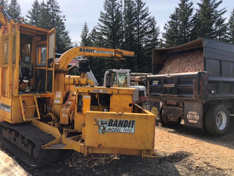 A wood chipper filling a dump truck with wood chips at a job site for Jere's Tree Service in Kodiak, AK.
