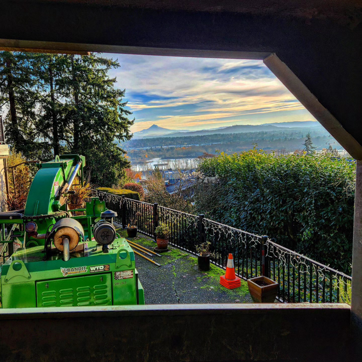 A green wood chipper at a Triple Acorn Tree Service job site in Portland, OR, with a scenic view of the city.