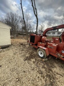 A wood chipper in a yard with wood chips and partially removed trees, showcasing tree removal and chipping services by Russell Tree Works & Firewood Sales in Augusta, ME.
