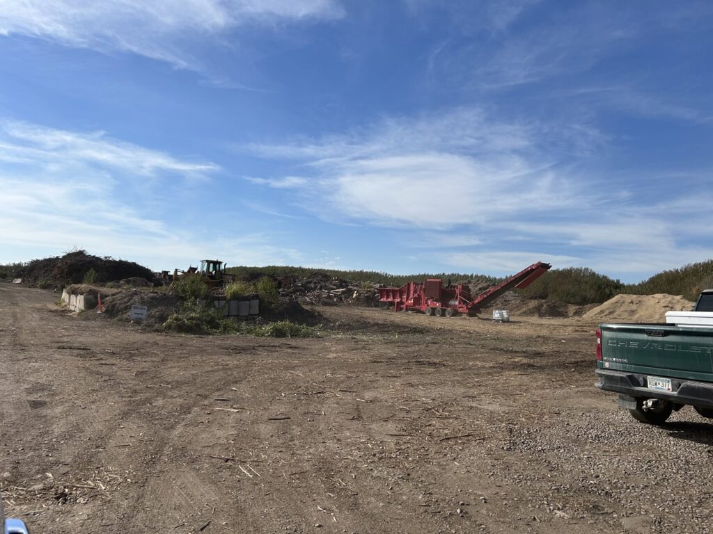 A wide view of the wood recycling site with a processing machine and piles of wood waste at Wood Recycling Site / Brush Dump in Rochester, MN