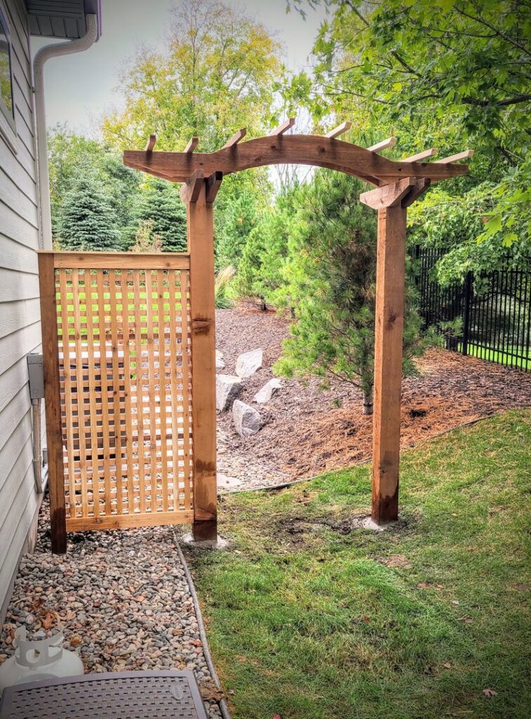 A decorative wooden archway or arbor with a lattice privacy screen, built by Minnesota Pergolas in Lakeland, MN.