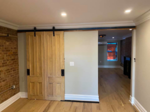 Stylish wooden barn door installed on a black track, enhancing a room with a brick accent wall by MTI Carpentry in Jersey City, NJ.