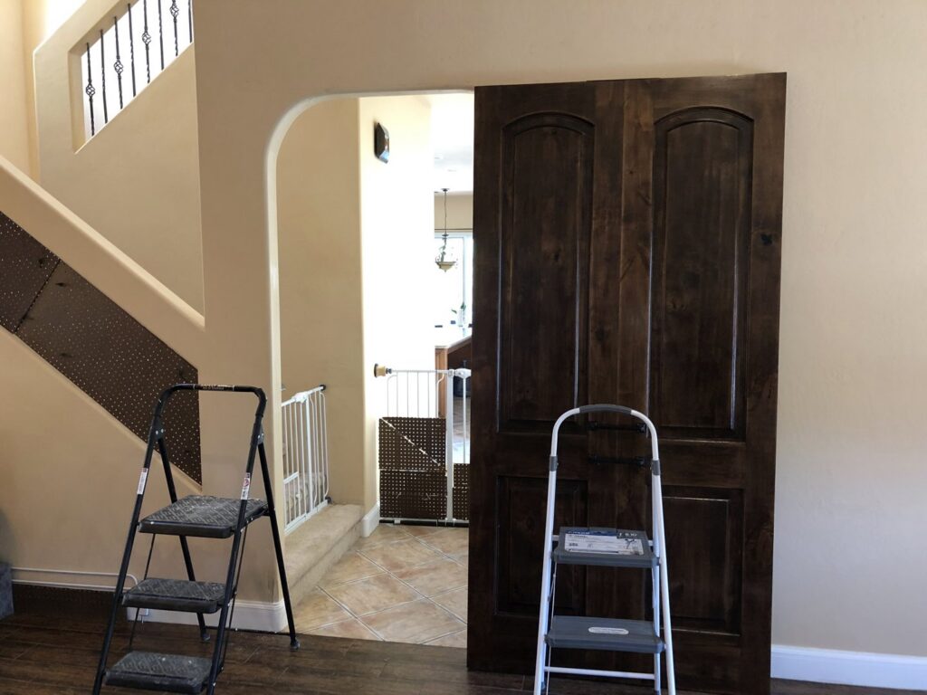 A large wooden door leaning against a wall with two step ladders, indicating a door installation or replacement project by The ABQ Handyman in Albuquerque, NM.