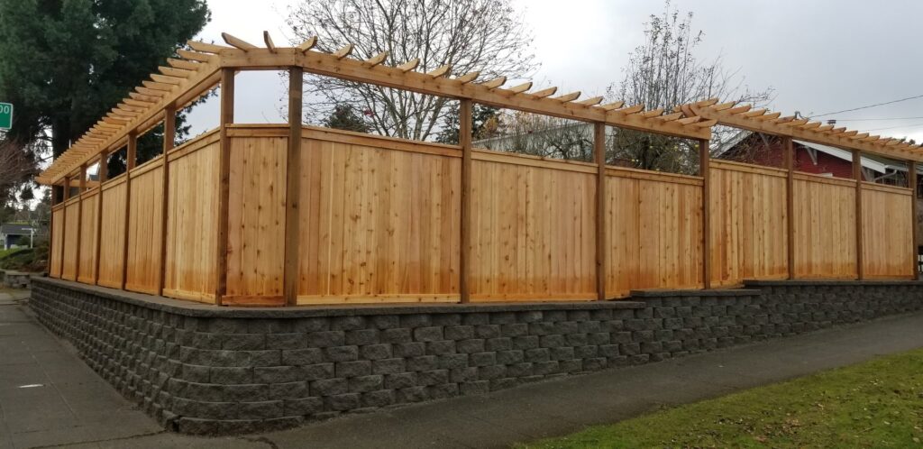 A custom wooden fence with a pergola-style top built on a stone retaining wall by LMS Landscapes & Fence services in Auburn, WA.