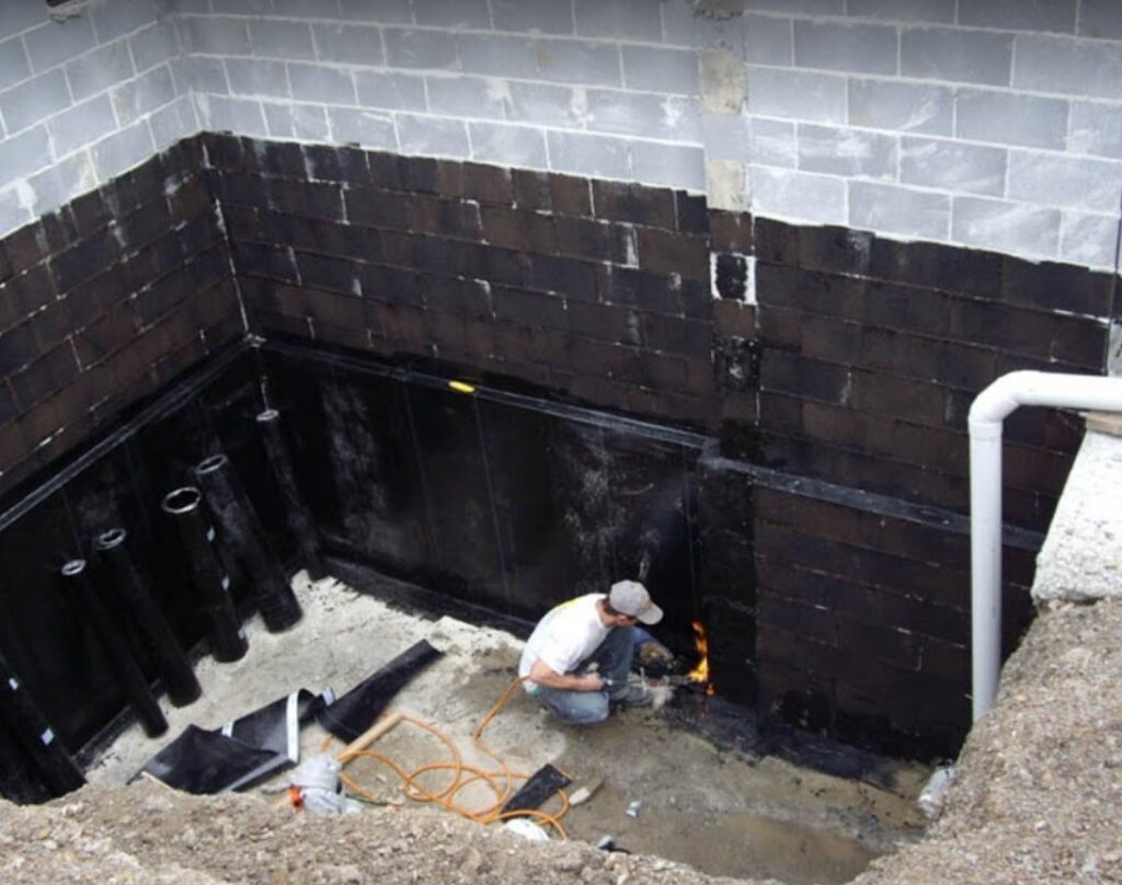 A worker applying waterproofing material to a basement wall, a service provided by Advanced Seamless Gutters, Inc. in Chicopee, MA.