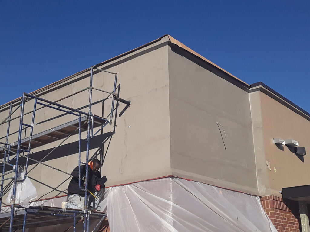 A worker on scaffolding applying material to a building corner, with protective sheeting, by Modern Wall Systems in Virginia Beach, VA.