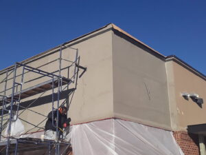 A worker on scaffolding applying material to a building corner, with protective sheeting, by Modern Wall Systems in Virginia Beach, VA.