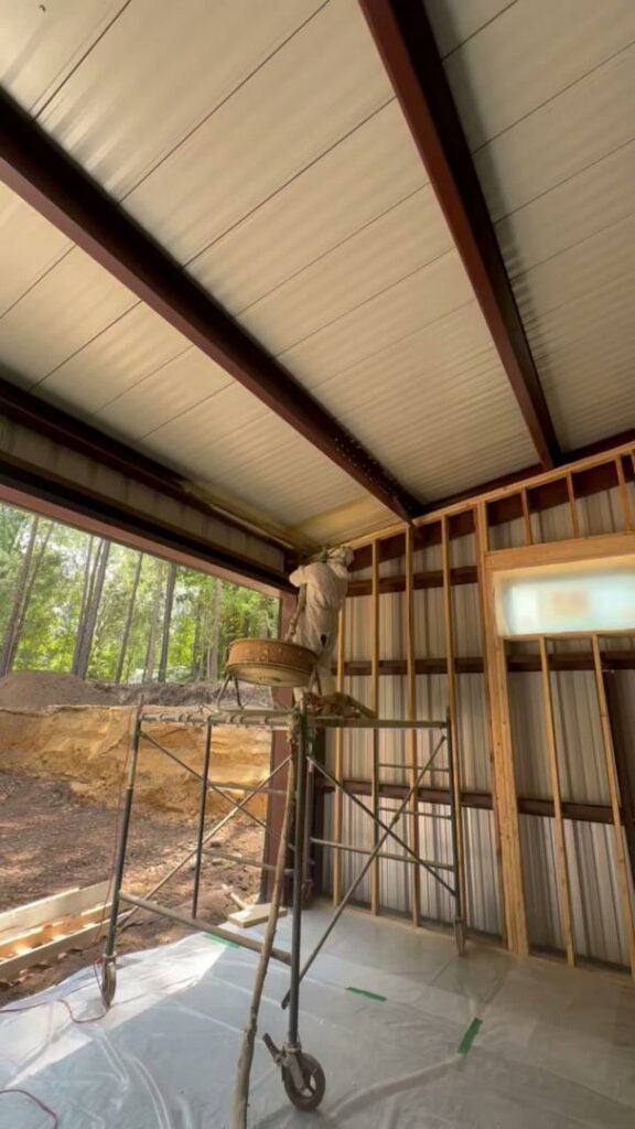 A worker on scaffolding applying spray foam insulation to a ceiling and wall by Koala Insulation of Charlotte, NC