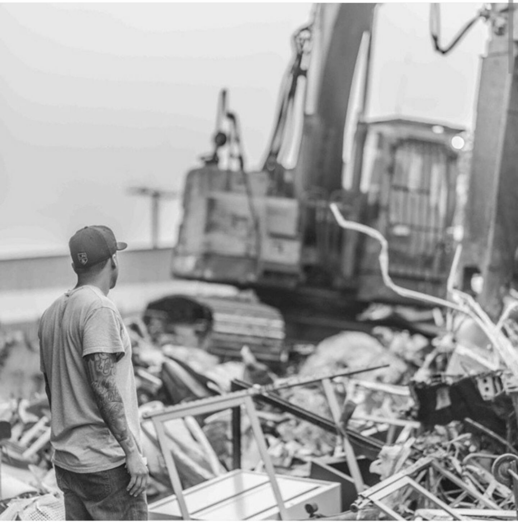 A worker stands near a large pile of mixed debris and an excavator, indicating a junk removal job by MIM Metal Recycling in Los Angeles, CA.