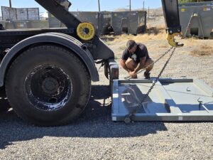 A Trash Panda worker attaching chains to equipment near a roll-off truck in Carson City, NV, for junk removal operations.