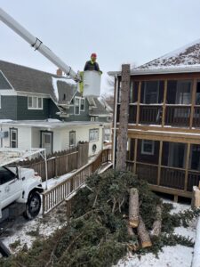 A worker in a bucket truck performing tree removal next to a house, with cut branches piled below, by Affordable Tree Service in Dickinson, ND.