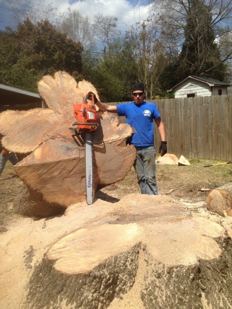 Worker with chainsaw next to large tree stump, showing tree felling or stump removal by Jon's Tree Service in Pelham, AL.